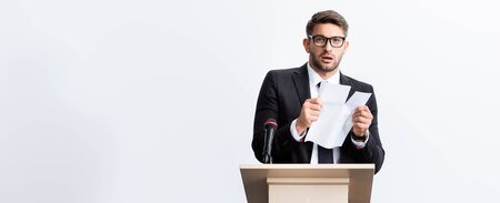 panoramic shot of scared businessman in suit standing at podium tribune and rearing paper during conference isolated on whiteの写真素材