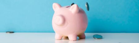 panoramic shot of falling silver coins near pink piggy bank on blue