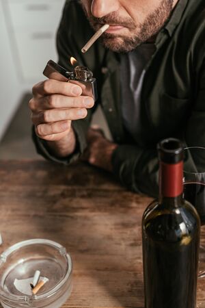 Cropped view of man lighting cigarette beside wine glass and bottle on tableの写真素材