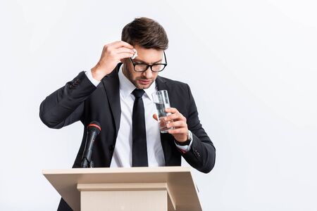 scared businessman in suit standing at podium tribune and holding glass of water during conference isolated on whiteの写真素材