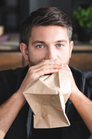 handsome man with panic attack breathing in paper bag in apartment
