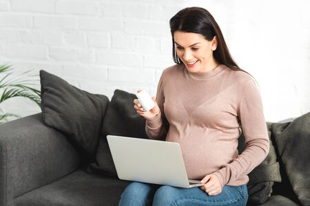 smiling pregnant woman holding pills and having online consultation with doctor on laptop at home