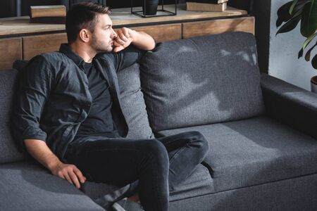 handsome and pensive man sitting on sofa and looking away in apartmentの写真素材