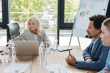 attractive, mature businesswoman sitting at desk near young multicultural colleagues during business...