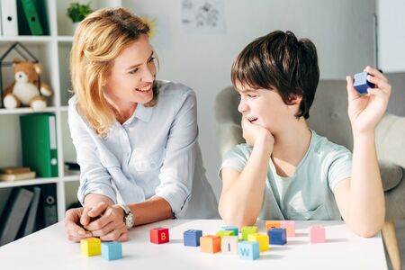 smiling child psychologist looking at kid with dyslexia and sitting at table with building blocksの写真素材