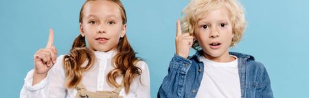 panoramic shot of shocked and cute kids looking at camera and showing idea sign isolated on blue