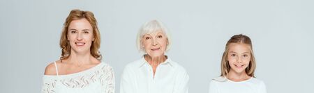 panoramic shot of smiling granddaughter, mother and grandmother looking at camera isolated on grey