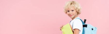 panoramic shot of smiling schoolboy with backpack holding book isolated on pink
