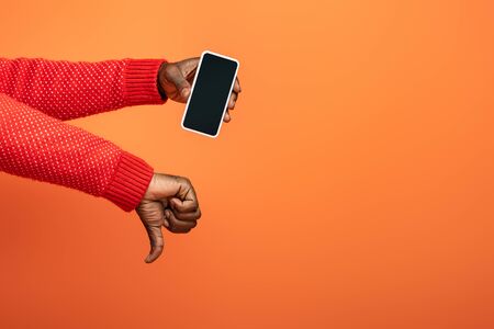 cropped view of african american man holding smartphone and showing thumb down on orange background