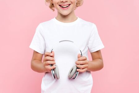 cropped view of smiling kid in t-shirt holding headphones isolated on pinkの写真素材