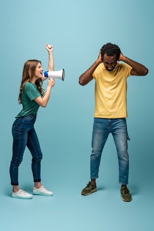 angry girl screaming in megaphone at african american boyfriend covering ears with hands on blue backgroundの写真素材
