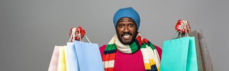 happy african american man in winter outfit with shopping bags on grey background, panoramic shot