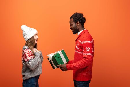side view of african american man gifting Christmas present to happy girlfriend on orange backgroundの写真素材