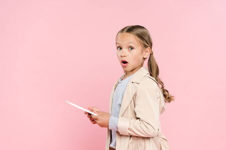 shocked kid holding digital tablet and looking at camera isolated on pinkの写真素材