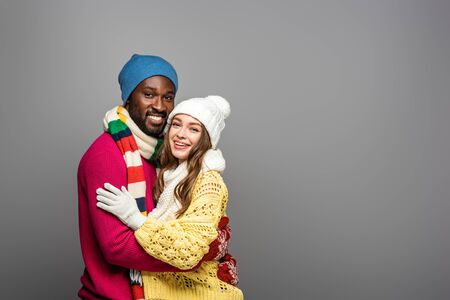 smiling interracial couple in winter outfit hugging on grey backgroundの写真素材