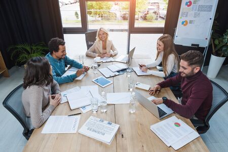 positive multicultural businesspeople sitting at wooden desk near digital devices, documents and...