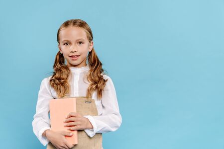 smiling and cute kid looking at camera and holding book isolated on blueの写真素材