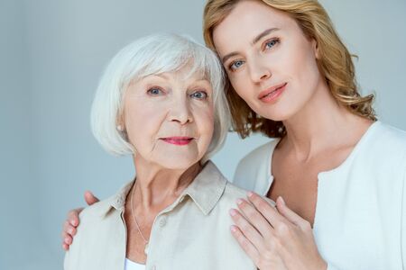daughter hugging smiling mother and looking at camera isolated on grey