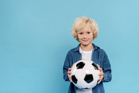smiling and cute kid looking at camera and holding football isolated on blueの写真素材