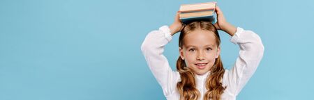 panoramic shot of smiling and cute kid looking at camera and holding books isolated on blueの写真素材