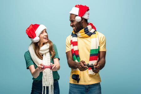 smiling interracial couple in santa hats and scarves holding joysticks isolated on blue