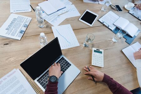 cropped view of businesspeople sitting at desk near digital devices, documents and glasses with...