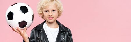 panoramic shot of kid in leather jacket holding football and looking at camera isolated on pinkの写真素材