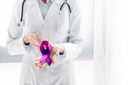 cropped view of doctor in white coat holding purple ribbon in clinic の写真素材