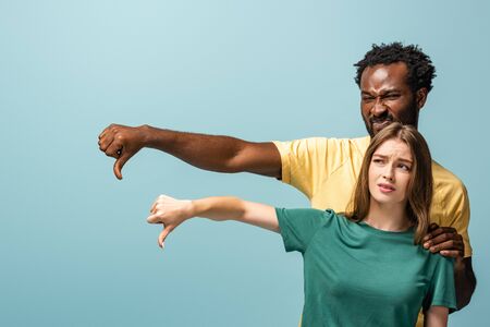 displeased interracial couple showing thumbs down isolated on blue backgroundの写真素材