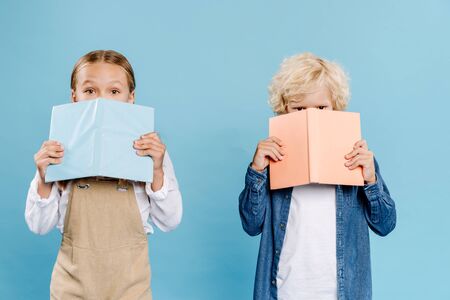kids looking at camera and obscuring faces with books isolated on blueの写真素材