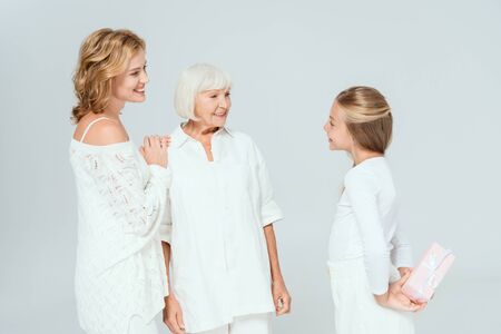 smiling granddaughter holding gift and looking at mother and grandmother isolated on grey