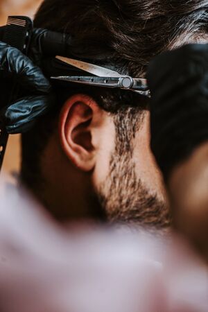 cropped view of barber in latex gloves holding hair comb and scissors while styling hair of man の写真素材