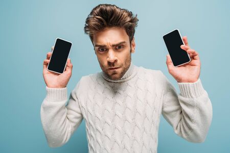 upset man showing two smartphones with blank screens, isolated on blue