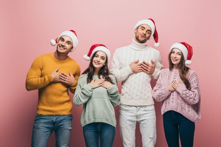 happy friends in sweaters and santa hats holding hands on hearts, isolated on pink