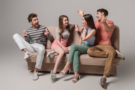 emotional young friends talking and sitting on sofa on grey