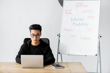 asian seo manager in glasses sitting near flipchart  with lettering internet marketing and using laptopの写真素材