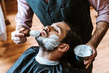 selective focus of barber applying shaving cream on face of man while holding bowl の写真素材