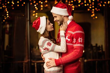 happy young couple in santa hats and warm sweaters embracing while looking at each otherの写真素材