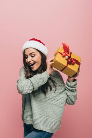 excited woman in sweater and santa hat holding christmas gift, isolated on pinkの写真素材