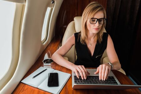 attractive businesswoman in glasses typing on laptop near smartphone with blank screen