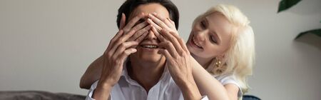 Smiling girl covering eyes to boyfriend at home, panoramic shot