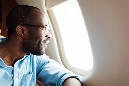 happy african american man in glasses looking at airplane window