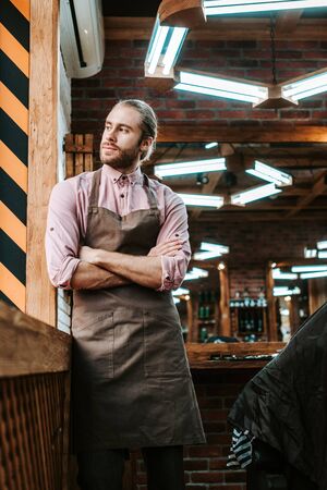 selective focus of handsome barber in apron standing with crossed arms and looking away