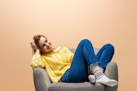 attractive smiling woman in yellow sweater sitting on armchair, isolated on beigeの写真素材