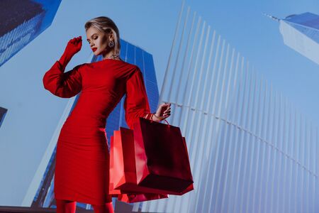 low angle view of attractive and stylish woman in red dress holding shopping bags on city backgroundの写真素材