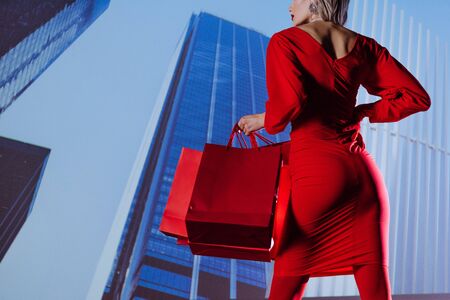 cropped view of stylish woman in red dress holding shopping bags on city background の写真素材