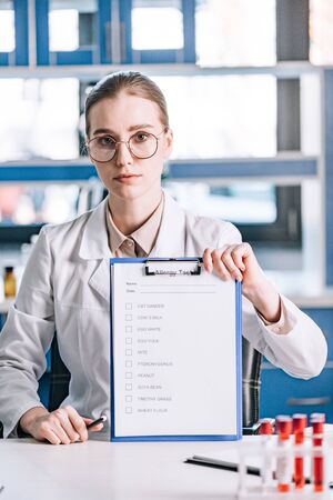 attractive immunologist holding clipboard with checklist and pen near test tubes