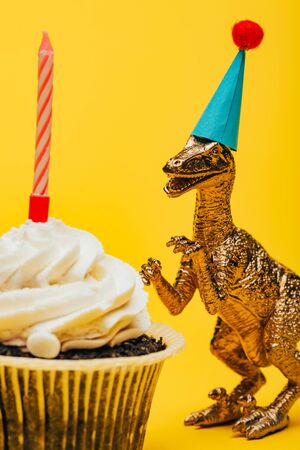 Selective focus of toy dinosaur in party cap beside cupcake with candle on yellow backgroundの写真素材