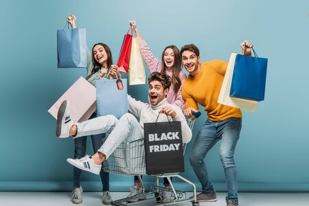 excited friends having fun with shopping bags in shopping cart on black friday, on blueの写真素材