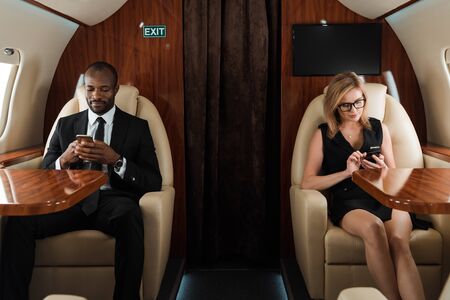 handsome african american businessman and attractive businesswoman using smartphones in private jetの写真素材
